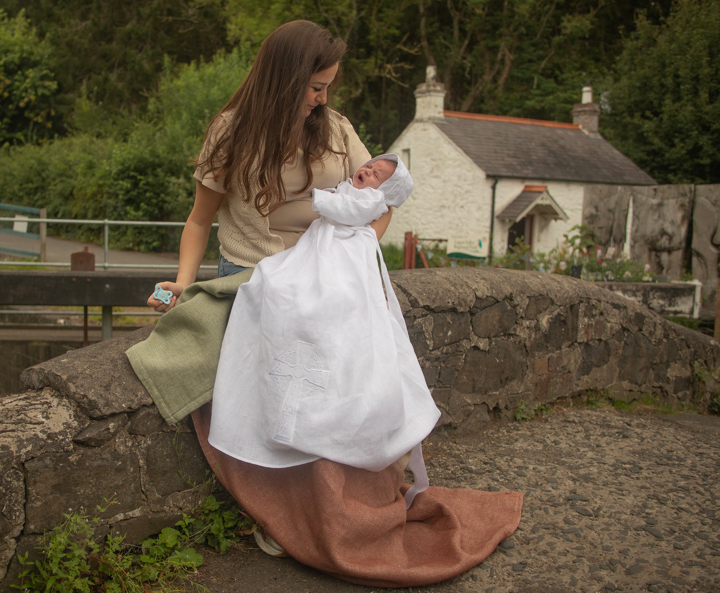 Mother holding a baby in teh Fionnuala christening gown by Brennans Irish Linens, on a stone bridge with a white cottage and trees in the background