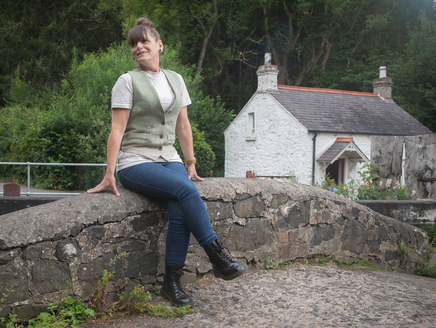 Woman sitting on a stone wall with a small cottage and greenery in the background
