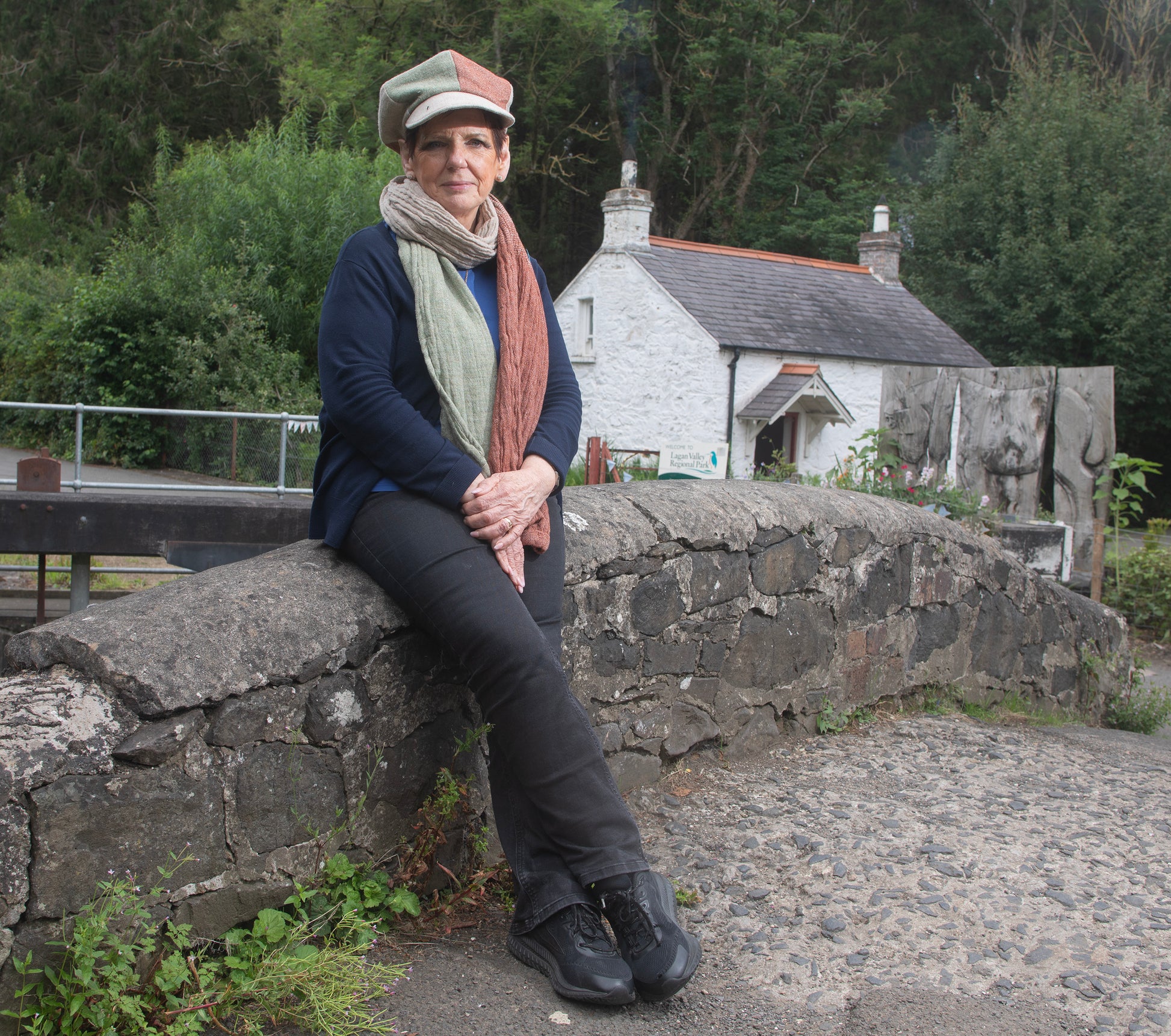 Eileen, the founder of Brennan's Irish Linens is wearing the Fae hat sitting on a bridge in front of a white cottage in Belfast