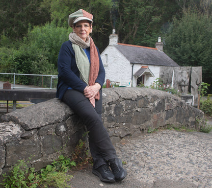 Eileen, the founder of Brennan's Irish Linens is wearing the Fae hat sitting on a bridge in front of a white cottage in Belfast
