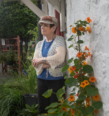 Eileen, the founder of Brennan's Irish Linens is wearing the Fae hat beside a white cottage with orange flowers beside her