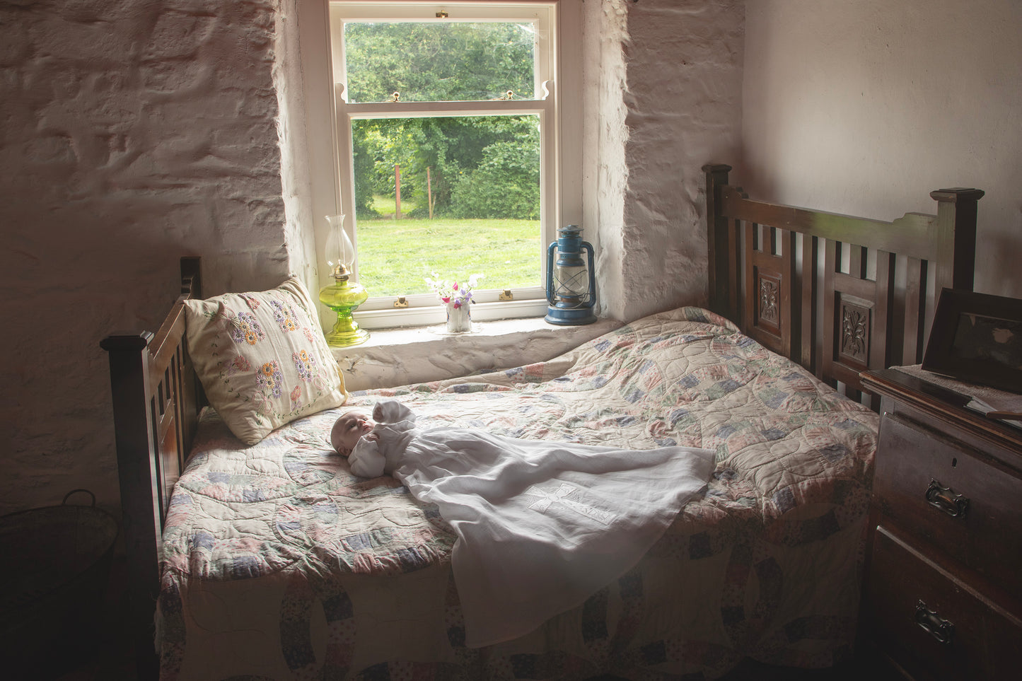 Bedroom in a cottage, with a baby wearing a christening gown on the bed, a window with greenery outside, and a dresser.