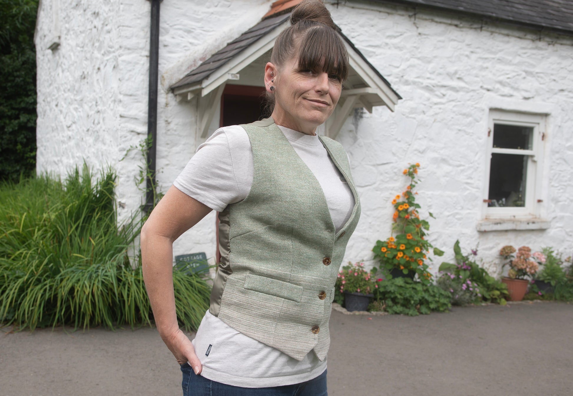 Woman wearing a green waistcoat over a white shirt in front of a white cottage with flowers around it.