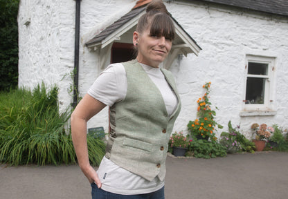 Woman wearing a green waistcoat over a white shirt in front of a white cottage with flowers around it.